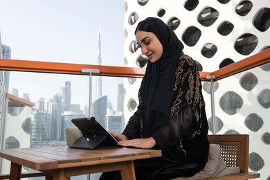 Side view of a woman reading from her tablet while standing in a modern cafe, a young veiled Arab Gulf Emirati woman wearing a black abaya embroidered with gold