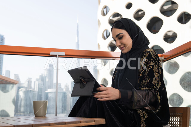 جلسة عمل هادئة مع إطلالة حضرية, احتراف الوظائف التقنية وتكنولوجيا المعلومات، متابعة آخر الأخبار على مواقع الانترنت، شابة عربية خليجية اماراتية محجبة ترتدي عباءة سوداء مطرزة باللون الذهبي