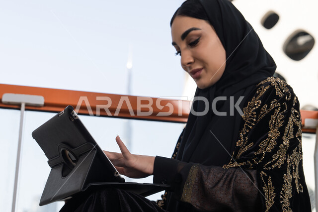 التواصل عن بعد واستخدام التطبيقات الاجتماعية على التابلت, صورة مقربة لامرأة عربية خليجية اماراتية تجلس في احدى المقاهي الفارهة و ترتدي عباءة سوداء اللون و مطرزة باللون الذهبي