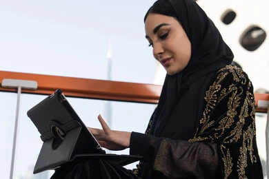 التواصل عن بعد واستخدام التطبيقات الاجتماعية على التابلت, صورة مقربة لامرأة عربية خليجية اماراتية تجلس في احدى المقاهي الفارهة و ترتدي عباءة سوداء اللون و مطرزة باللون الذهبي
