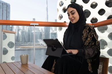 Following the latest news on the Internet, a young veiled Emirati Gulf Arab woman wearing a black abaya embroidered with gold, and next to her is a cup of hot drink, using a tablet device