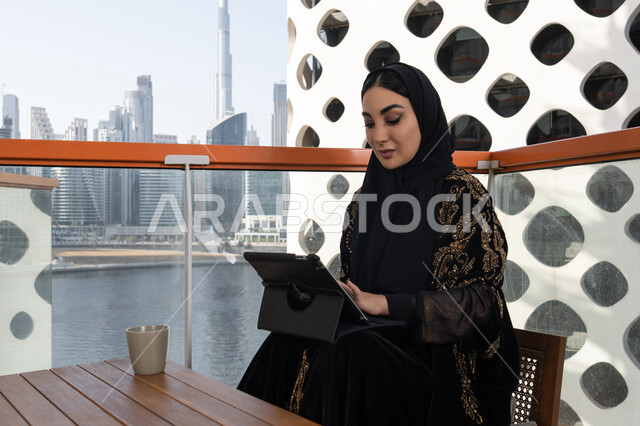 ميزات تقنية حديثة ومتطورة, إدارة المشاريع عن بعد, شابة عربية خليجية اماراتية محجبة ترتدي عباءة سوداء مطرزة باللون الذهبي وبجانبها كوب من المشروب الدافئ, استخدام الجهاز لوحي