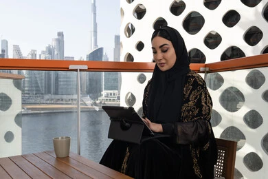 ميزات تقنية حديثة ومتطورة, إدارة المشاريع عن بعد, شابة عربية خليجية اماراتية محجبة ترتدي عباءة سوداء مطرزة باللون الذهبي وبجانبها كوب من المشروب الدافئ, استخدام الجهاز لوحي