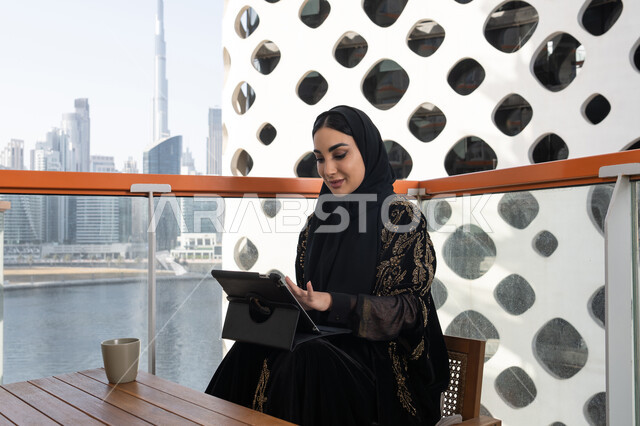 Remote project management, a young veiled Emirati Gulf Arab woman wearing a black abaya embroidered with gold, and next to her is a cup of hot drink, using a tablet