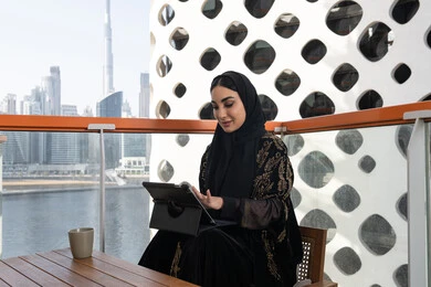 إدارة المشاريع عن بعد, شابة عربية خليجية اماراتية محجبة ترتدي عباءة سوداء مطرزة باللون الذهبي وبجانبها كوب من المشروب الدافئ, استخدام الجهاز لوحي