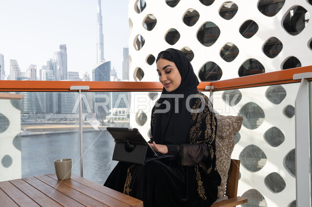 إنجاز مهام العمل عبر التابلت, شابة عربية خليجية اماراتية محجبة ترتدي عباءة سوداء مطرزة باللون الذهبي و بجانبها كوب من المشروب الدافئ, استخدام الجهاز اللوحي