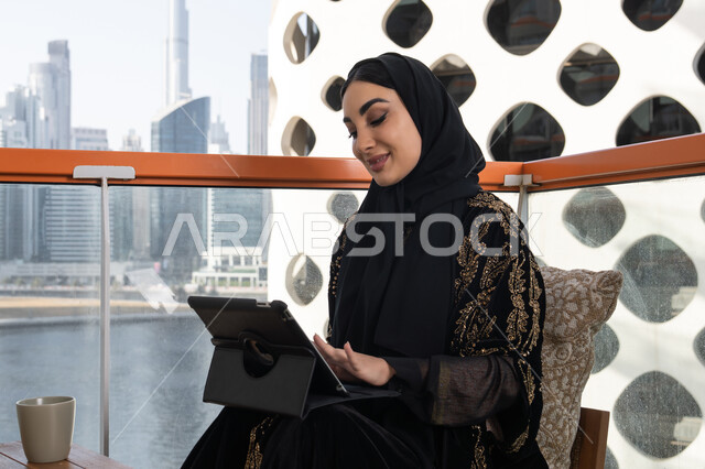 القيام بمقابلات وإجتماعات العمل أونلاين، شابة عربية خليجية اماراتية محجبة ترتدي عباءة سوداء مطرزة باللون الذهبي وبجانبها كوب من المشروب الدافئ, استخدام الجهاز لوحي