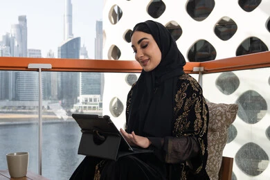 القيام بمقابلات وإجتماعات العمل أونلاين، شابة عربية خليجية اماراتية محجبة ترتدي عباءة سوداء مطرزة باللون الذهبي وبجانبها كوب من المشروب الدافئ, استخدام الجهاز لوحي