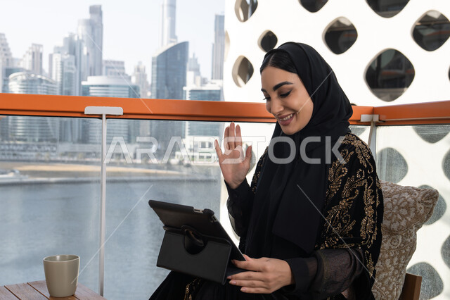 جلسة عمل عن بعد، إجراء مكالمات الفيديو باستخدام جهاز التابلت، شابة عربية خليجية اماراتية محجبة ترتدي عباءة سوداء مطرزة باللون الذهبي وبجانبها كوب من المشروب الدافئ, تعابير إلقاء السلام والتحية