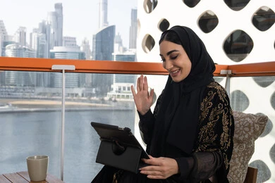 جلسة عمل عن بعد، إجراء مكالمات الفيديو باستخدام جهاز التابلت، شابة عربية خليجية اماراتية محجبة ترتدي عباءة سوداء مطرزة باللون الذهبي وبجانبها كوب من المشروب الدافئ, تعابير إلقاء السلام والتحية