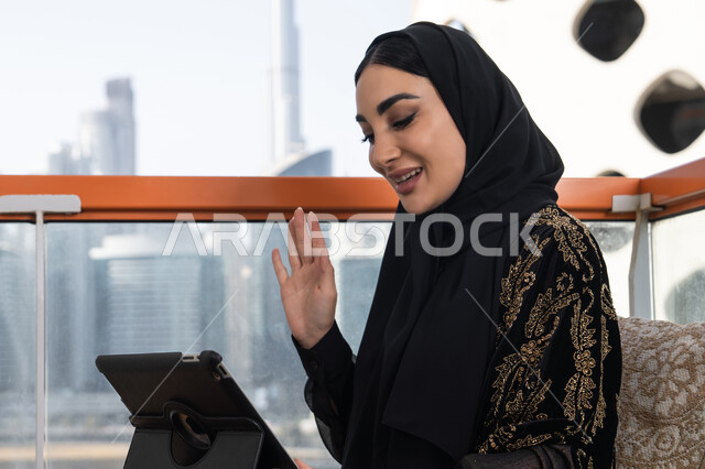 جلسة معاصرة هادئة, إجراء مكالمات الفيديو باستخدام جهاز التابلت، شابة عربية خليجية اماراتية محجبة ترتدي عباءة سوداء مطرزة باللون الذهبي, تعابير إلقاء السلام والتحية