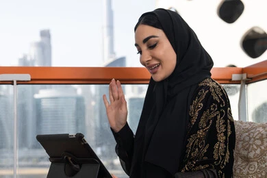 جلسة معاصرة هادئة, إجراء مكالمات الفيديو باستخدام جهاز التابلت، شابة عربية خليجية اماراتية محجبة ترتدي عباءة سوداء مطرزة باللون الذهبي, تعابير إلقاء السلام والتحية