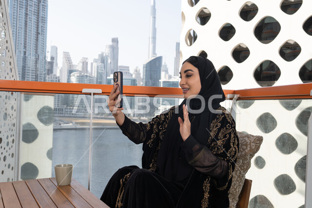 إجراء مكالمات الفيديو عبر الموبايل، تعابير إلقاء السلام والتحية, استخدام جهاز تقني حديث ومتطور, التواصل مع الأصدقاء والعائلة, شابة عربية خليجية اماراتية محجبة ترتدي عباءة سوداء مطرزة باللون الذهبي  تمسك بيدها هاتف محمول و بجانبها كوب من المشروب الدافئ