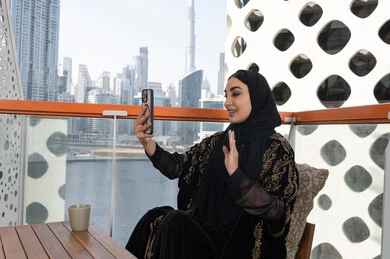 إجراء مكالمات الفيديو عبر الموبايل، تعابير إلقاء السلام والتحية, استخدام جهاز تقني حديث ومتطور, التواصل مع الأصدقاء والعائلة, شابة عربية خليجية اماراتية محجبة ترتدي عباءة سوداء مطرزة باللون الذهبي  تمسك بيدها هاتف محمول و بجانبها كوب من المشروب الدافئ