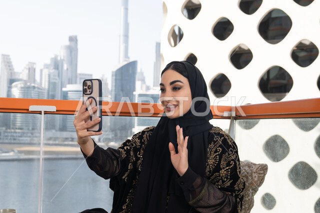 Expressions of greeting and peace, using a modern and advanced technical device, communicating with friends and family, a young veiled Emirati Gulf Arab woman wearing a black abaya embroidered in gold and holding a mobile phone in her hand