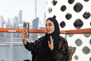 تعابير إلقاء السلام والتحية, استخدام جهاز تقني حديث ومتطور, التواصل مع الأصدقاء والعائلة, شابة عربية خليجية اماراتية محجبة ترتدي عباءة سوداء مطرزة باللون الذهبي و تمسك بيدها هاتف محمول