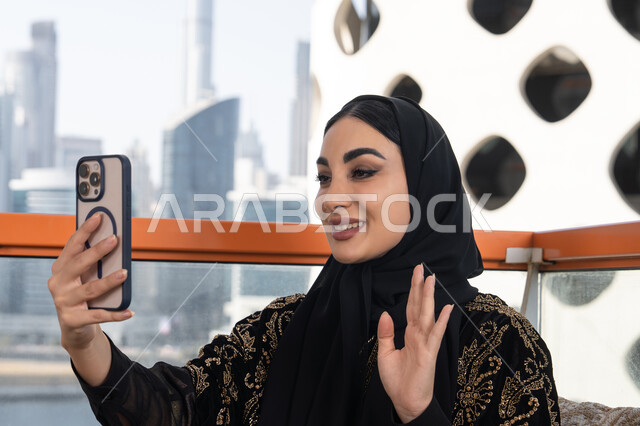إجراء مكالمة فيديو بتعابير الفرح والسرور, دردشة و اتصالات مع الأهل والأصدقاء, جلسة عصرية في مقهى حديث وإطلالة ناطحات السحاب, شابة عربية خليجية اماراتية محجبة ترتدي عباءة سوداء مطرزة باللون الذهبي و تمسك بيدها هاتف محمول