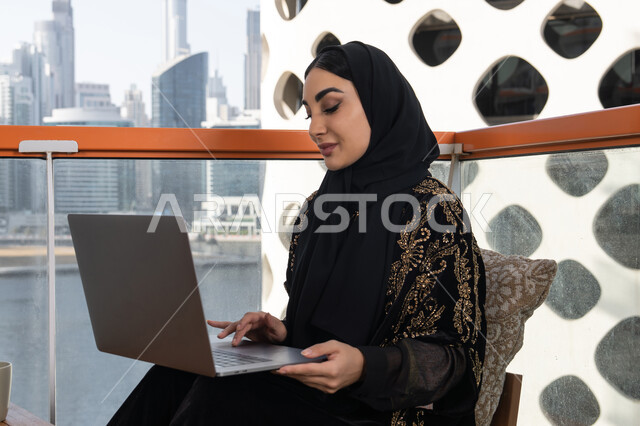 جلسة عصرية في مقهى حديث وإطلالة ناطحات السحاب, شابة عربية خليجية اماراتية محجبة  ترتدي عباءة سوداء اللون, العمل على جهاز الكمبيوتر المحمول