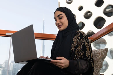 جلسة معاصرة هادئة, شابة عربية خليجية اماراتية محجبة ترتدي عباءة سوداء مطرزة باللون الذهبي, العمل على جهاز الكمبيوتر المحمول