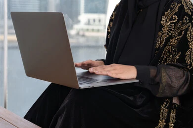 صورة مقربة لامرأة عربية خليجية اماراتية تعمل على اللابتوب في احدى المقاهي الفارهة و ترتدي عباءة سوداء اللون و مطرزة باللون الذهبي, العمل على جهاز الكمبيوتر المحمول