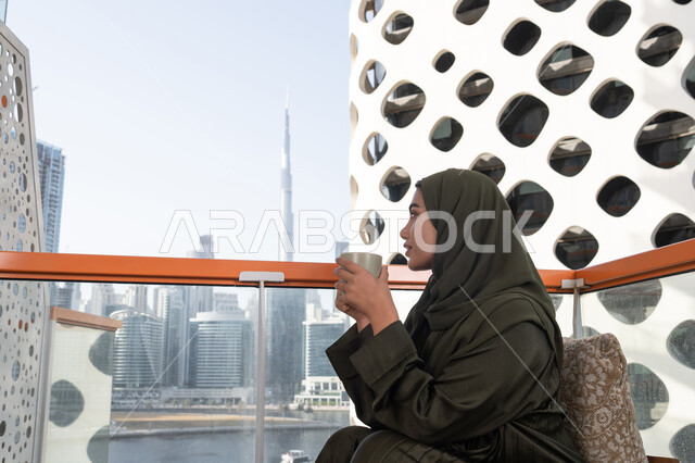 An elegant view from a high-rise cafe overlooking the city skyline, a young veiled Emirati Gulf Arab woman wearing a green abaya and holding a cup of hot drink, a warm and calm atmosphere