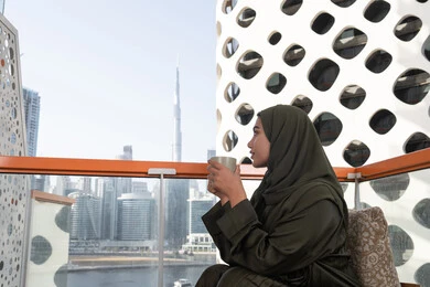 إطلالة أنيقة من مقهى مرتفع يطل على أفق المدينة, شابة عربية خليجية اماراتية محجبة و ترتدي عباءة خضراء و تحمل كوب من المشروب الساخن, أجواء دافئة هادئة