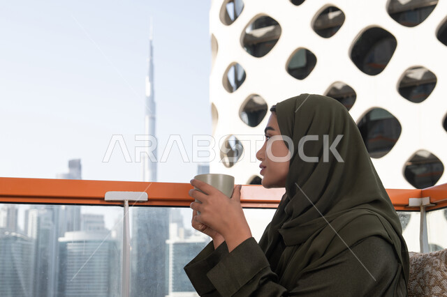 مقهى راقي بالامارات، إطلالة أنيقة من مقهى مرتفع يطل على أفق المدينة, شابة عربية خليجية اماراتية محجبة ترتدي عباءة خضراء و حجاب و تحمل كوب من المشروب الساخن, أجواء دافئة هادئة