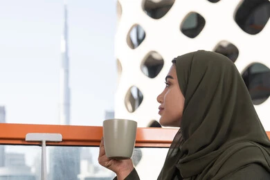 أجواء دافئة هادئة, صورة مقربة لامرأة تنظر بعيدا من الأعلى في المقهى, شابة عربية خليجية اماراتية محجبة ترتدي عباءة خضراء و حجاب و تحمل كوب من المشروب الساخن