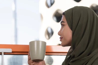 صورة مقربة لامرأة تنظر بعيدا من الأعلى في المقهى, شابة عربية خليجية اماراتية محجبة ترتدي عباءة خضراء و حجاب و تحمل كوب من المشروب الساخن, أجواء دافئة هادئة, تسلية وترفيه في احدى المقاهي المطلة على المدينة