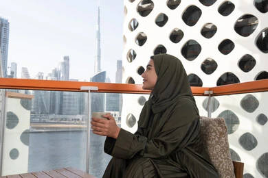 تسلية وترفيه في احدى المقاهي المطلة على المدينة, شابة عربية خليجية اماراتية محجبة ترتدي عباءة خضراء و حجاب و تحمل كوب من المشروب الساخن, أجواء دافئة هادئة