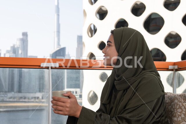 Gestures of relaxation and tranquility, one of the upscale cafes in the Emirates, an elegant view from a high cafe overlooking the city skyline, a veiled young Emirati Gulf Arab woman wearing a green abaya and hijab and holding a cup of hot drink, a warm and calm atmosphere