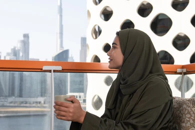 ايماءات الاستجمام والسكينة, احدى المقاهي الراقية بالامارات، إطلالة أنيقة من مقهى مرتفع يطل على أفق المدينة, شابة عربية خليجية اماراتية محجبة ترتدي عباءة خضراء و حجاب و تحمل كوب من المشروب الساخن, أجواء دافئة هادئة