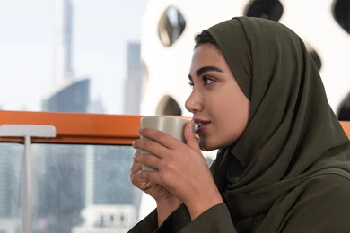 امرأة تمسك بيدها كوب من المشروب الدافئ, صورة مقربة لامرأة تنظر بعيدا من الأعلى في المقهى, شابة عربية خليجية اماراتية محجبة ترتدي عباءة خضراء و حجاب, أجواء دافئة هادئة