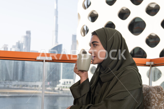 Drinking coffee and hot drinks, enjoying coffee in a quiet atmosphere, a veiled Emirati Gulf Arab woman wearing a green abaya sitting in a high cafe overlooking the city