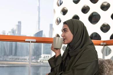 تناول القهوة والمشروبات الساخنة، الاستمتاع بشرب القهوة في أجواء هادئة، امرأة عربية خليجية اماراتية محجبة ترتدي عباءة خضراء تجلس في مقهى مرتفع مطل على المدينة