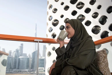 الاستمتاع بشرب القهوة في أجواء هادئة، امرأة عربية خليجية اماراتية محجبة ترتدي عباءة خضراء تجلس في مقهى مرتفع مطل على المدينة