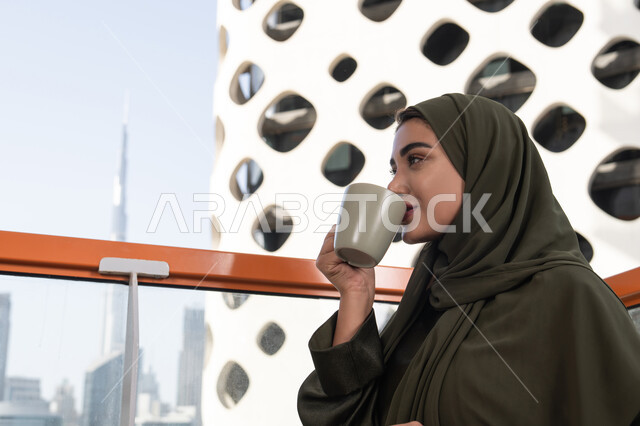 شرب مشروب ساخن ولذيذ, مفهوم الإستمتاع بالمشروبات الدافئة, امرأة عربية خليجية اماراتية محجبة ترتدي عباءة خضراء تجلس في مقهى مرتفع مطل على المدينة