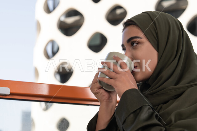 قضاء الوقت باحتساء المشروب المفضل, مفهوم الإستمتاع بالمشروبات الدافئة, امرأة عربية خليجية اماراتية محجبة ترتدي عباءة خضراء تجلس في مقهى مرتفع مطل على المدينة