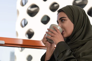 قضاء الوقت باحتساء المشروب المفضل, مفهوم الإستمتاع بالمشروبات الدافئة, امرأة عربية خليجية اماراتية محجبة ترتدي عباءة خضراء تجلس في مقهى مرتفع مطل على المدينة