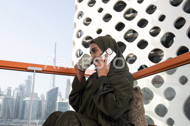 مفهوم الإستمتاع بالمشروبات الدافئة, امرأة عربية خليجية اماراتية محجبة ترتدي عباءة خضراء تقوم بإجراء مكالمة هاتفية في مقهى في الهواء الطلق, استخدام الأجهزة والتقنيات الحديثة والمتطورة