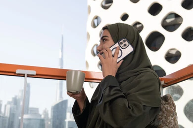 التواصل والاطمئنان على الأصدقاء, الاستمتاع بتناول المشروب الساخن, امرأة عربية خليجية اماراتية محجبة ترتدي عباءة خضراء تقوم بإجراء مكالمة هاتفية في مقهى فاخر مرتفع مطل على المدينة
