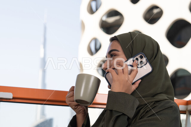 مفهوم صلة الرحم، محادثات ودردشات مع الاصدقاء، الاستمتاع بتناول المشروب الساخن, امرأة عربية خليجية اماراتية محجبة ترتدي عباءة خضراء تقوم بإجراء مكالمة هاتفية في مقهى في الهواء الطلق