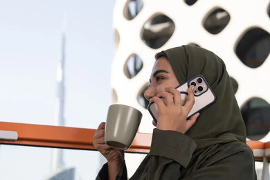 مفهوم صلة الرحم، محادثات ودردشات مع الاصدقاء، الاستمتاع بتناول المشروب الساخن, امرأة عربية خليجية اماراتية محجبة ترتدي عباءة خضراء تقوم بإجراء مكالمة هاتفية في مقهى في الهواء الطلق