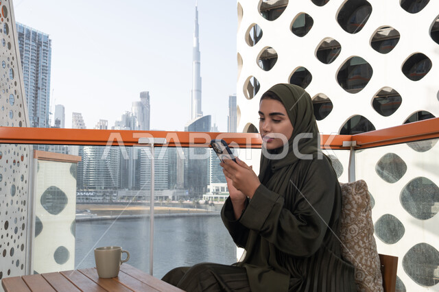 Using modern technology for entertainment and fun, spending enjoyable times in an outdoor cafe, a young veiled Emirati Gulf Arab woman wearing a green abaya holding a mobile phone and a hot drink next to her