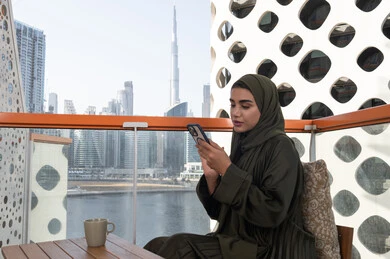 استخدام الأجهزة التقنية الحديثة للتسلية و الترفيه, قضاء اوقات ممتعة في مقهى في الهواء الطلق،  شابة عربية خليجية اماراتية محجبة ترتدي عباءة خضراء تحمل هاتف محمول و بجانبها مشروب ساخن