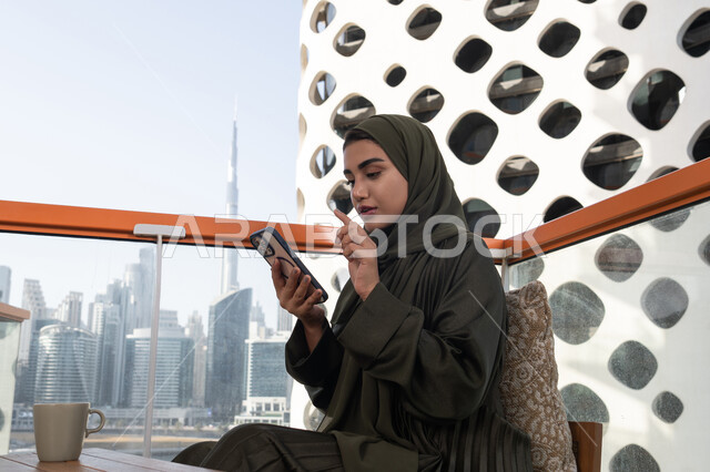 تصفح تطبيقات المحمول، النقر على شاشة الهاتف الخلوي, شابة عربية خليجية اماراتية محجبة ترتدي عباءة خضراء تجلس في مقهى و بجانبها مشروب ساخن, استخدام التقنيات الحديثة والمتطورة