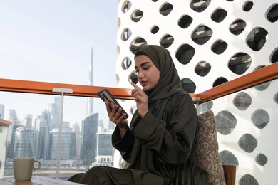 تصفح تطبيقات المحمول، النقر على شاشة الهاتف الخلوي, شابة عربية خليجية اماراتية محجبة ترتدي عباءة خضراء تجلس في مقهى و بجانبها مشروب ساخن, استخدام التقنيات الحديثة والمتطورة