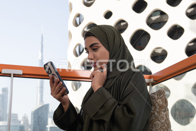 إيماءات وجه ويدين تعبر عن الحيرة, إستخدام اجهزة تقنية حديثة، تصفح وسائل التواصل الاجتماعي عبر الجوال, سيدة عربية خليجية اماراتية ترتدي الحجاب مع عباءة خضراء تجلس في مقهى في الهواء الطلق