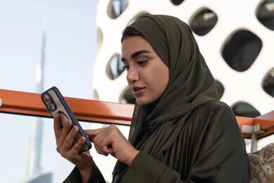 تصفح وسائل التواصل الاجتماعي عبر الجوال, سيدة عربية خليجية اماراتية ترتدي الحجاب مع عباءة خضراء تجلس في مقهى في الهواء الطلق,  استخدام الأجهزة التقنية الحديثة