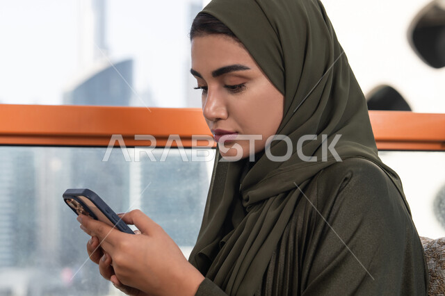 النقر على شاشة الهاتف المحمول, تصفح وسائل التواصل الاجتماعي عبر الجوال, سيدة عربية خليجية اماراتية ترتدي الحجاب مع عباءة خضراء تجلس في مقهى في الهواء الطلق, استخدام الأجهزة التقنية الحديثة